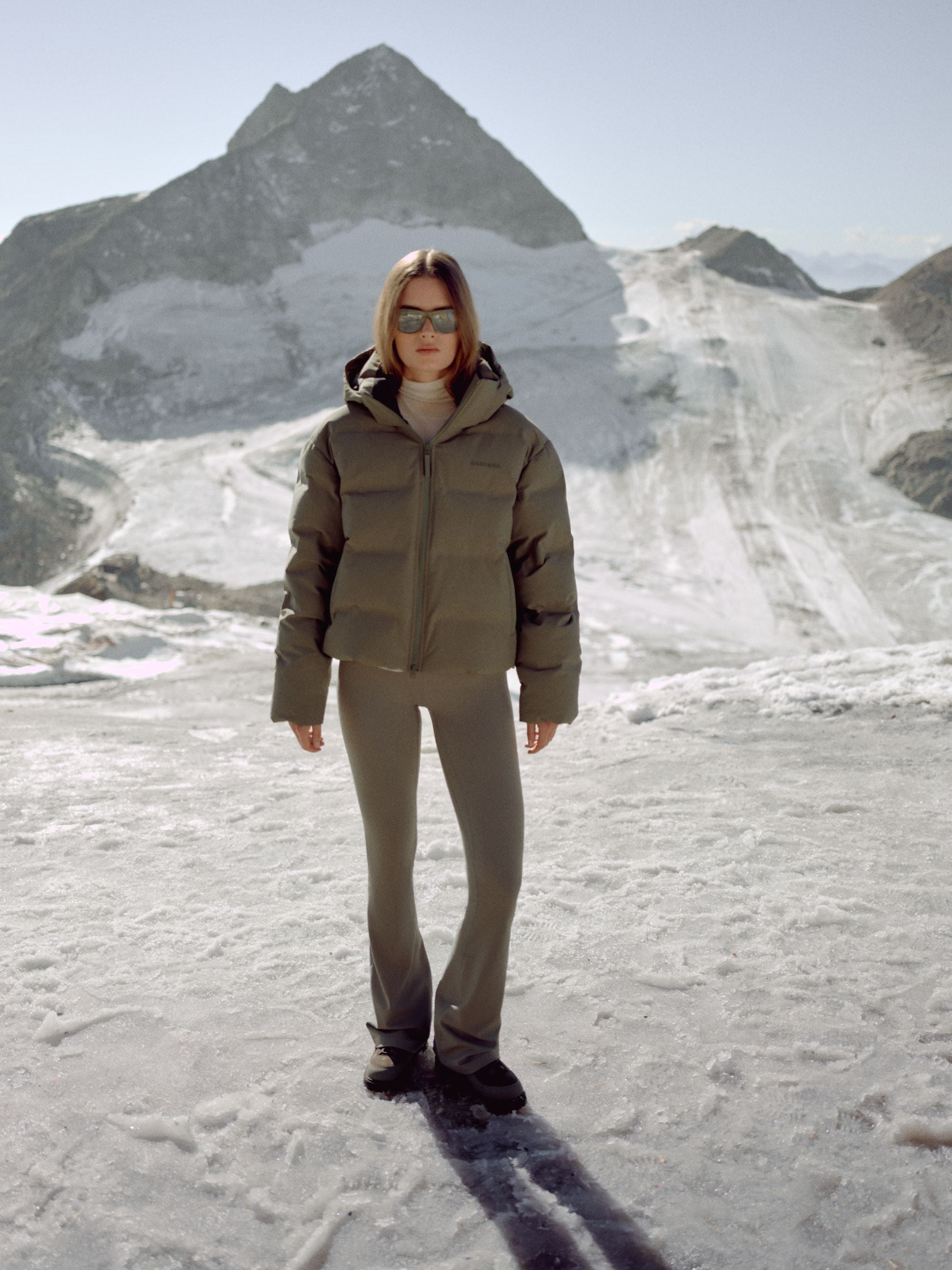 Woman wearing a khaki CASPARA Aurora puffer jacket standing in a snowy mountain landscape.